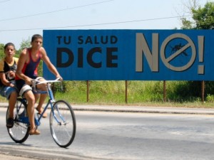 Cubanos en bicicleta pasan por delante a una propaganda de la campaña para erradicar el mosquito que transmite el dengue hemorrágico.