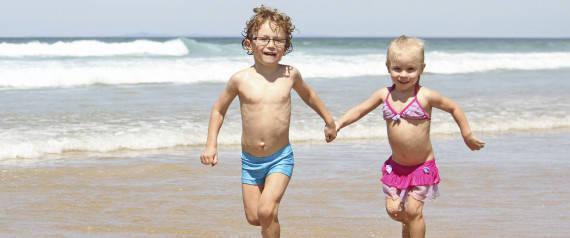 A little boy and girl, brother & sister, running together in the water at the beach, wearing swimmers, having fun.