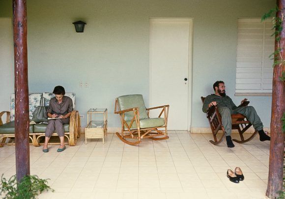Fidel descansando en el portal de una casa en la Isla de Pinos (hoy Isla de la Juventud). En el otro extremo de la foto, Celia Sánchez hace apuntes. 1965. Foto: Lee Lockwood